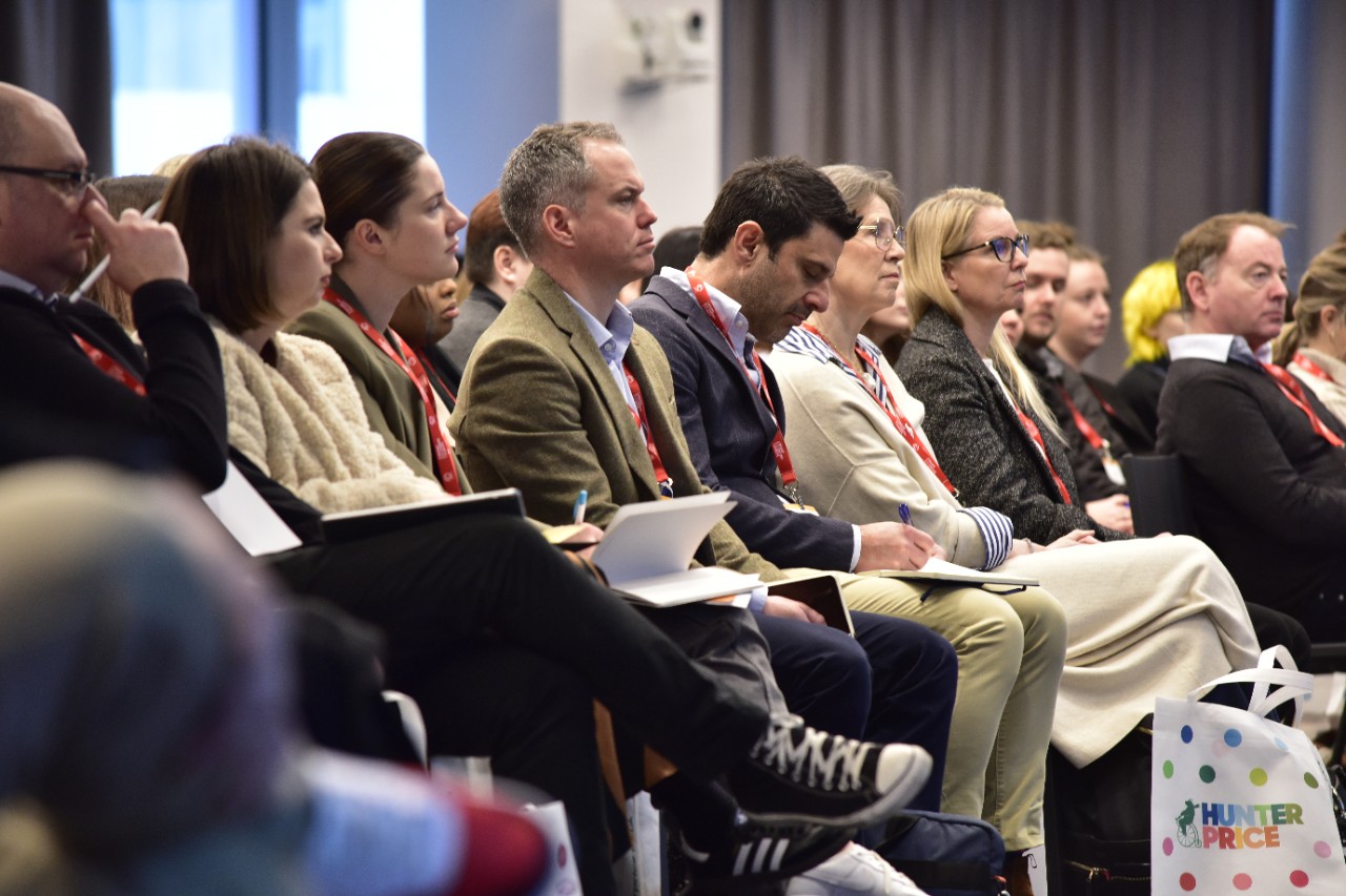 Retail professionals attending a brand licensing conference, listening to a speaker and taking notes. Attendees wear red lanyards, signifying participation in an industry event focused on retail strategies and brand partnerships. A Hunter Price shopping bag is visible, highlighting retail collaborations.