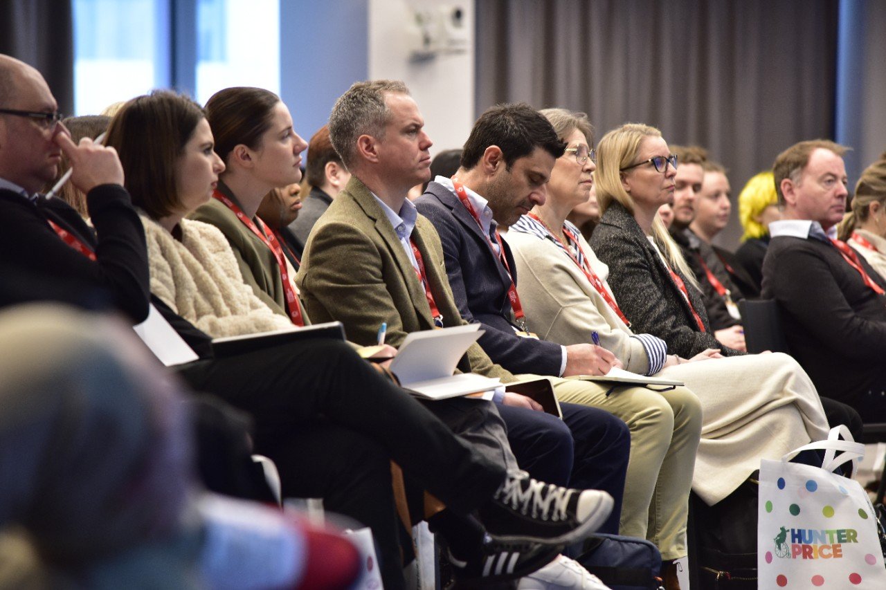 Retail professionals attending a brand licensing conference, listening to a speaker and taking notes. Attendees wear red lanyards, signifying participation in an industry event focused on retail strategies and brand partnerships. A Hunter Price shopping bag is visible, highlighting retail collaborations.