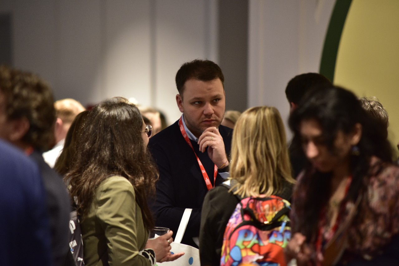 Retail and brand licensing professionals networking at an industry event. A man wearing a red lanyard listens intently while engaging in discussions with attendees. The setting highlights business collaboration and partnership opportunities in the licensing sector.