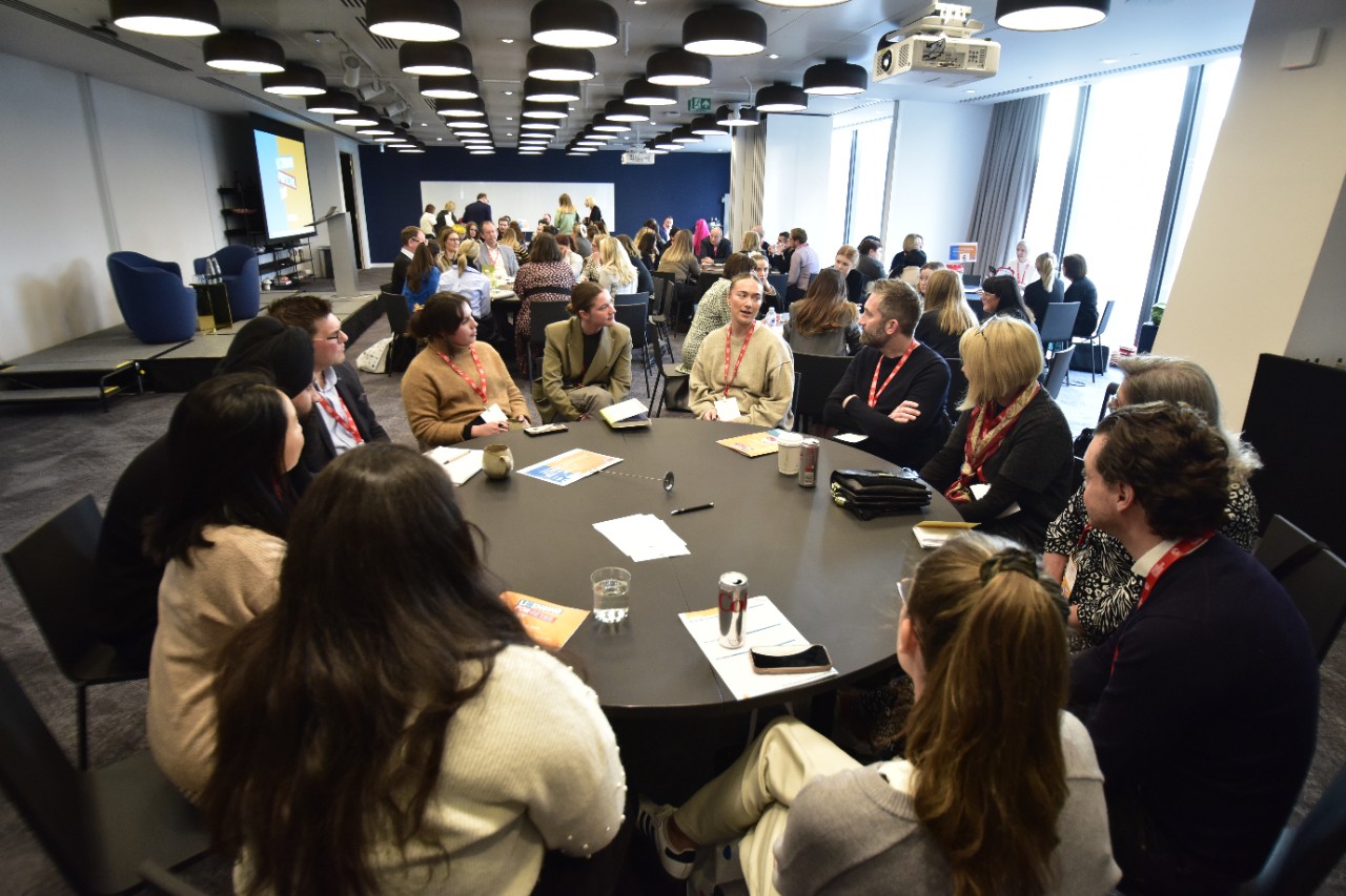 Retail professionals participating in a roundtable discussion at a brand licensing networking event. Attendees wearing red lanyards engage in conversations about how brand licensing strategies can drive business growth. The session takes place in a modern conference room with a presentation screen in the background.