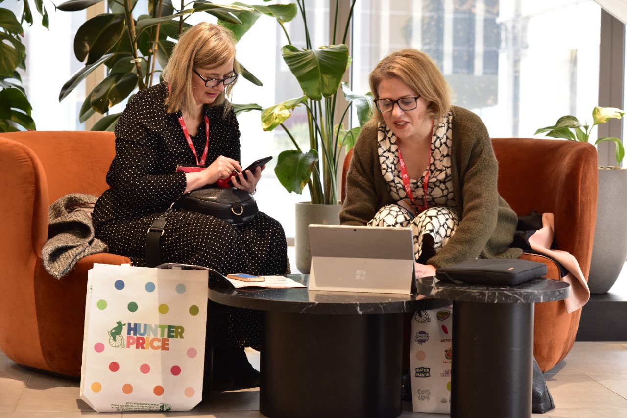 Two retail professionals at a brand licensing event collaborating on a laptop. They wear red lanyards and sit in a modern lounge area, discussing business opportunities. A Hunter Price shopping bag and other branded materials are visible, highlighting retail partnerships."